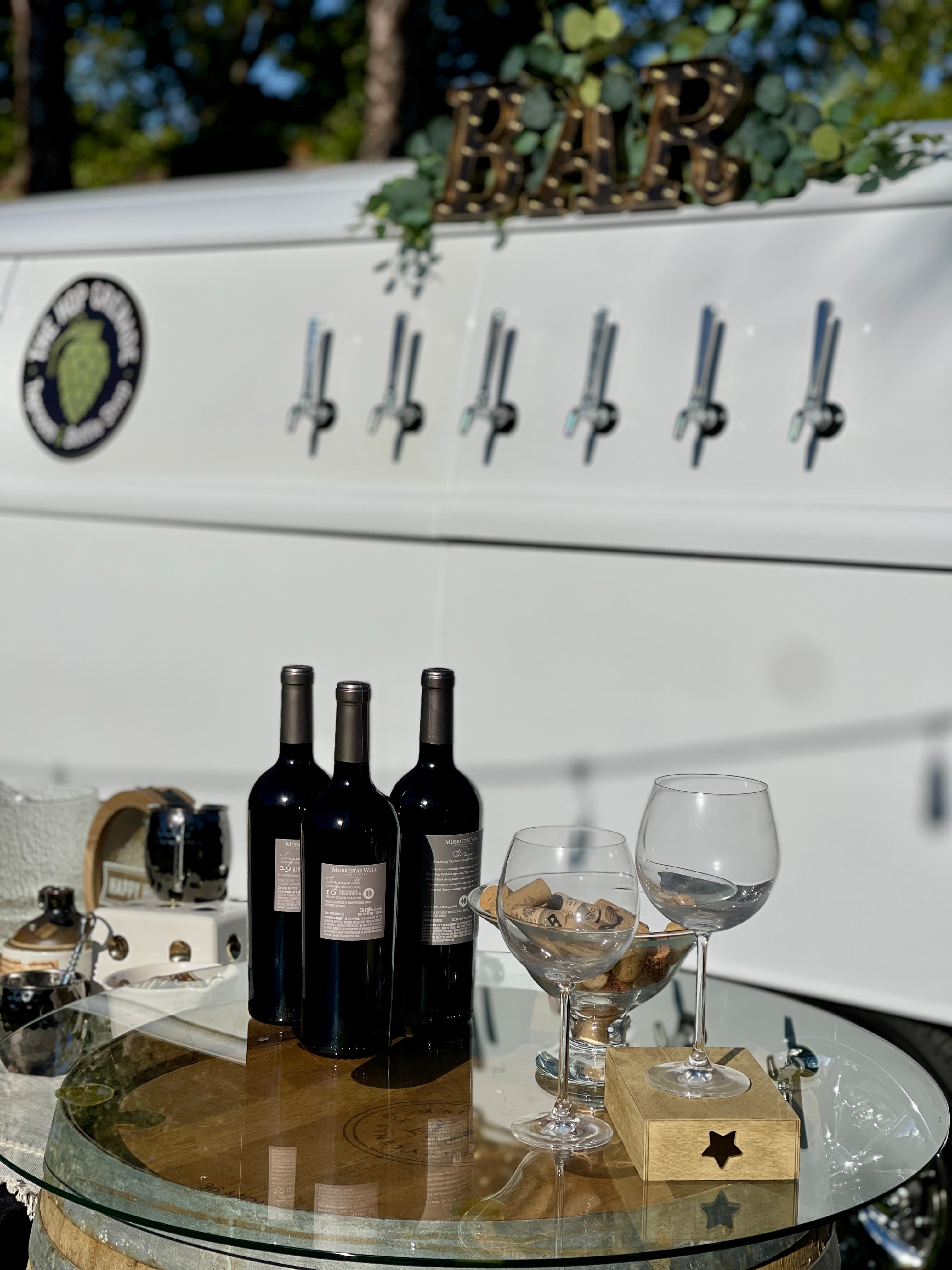 Three wine bottles and glasses on a barrel table before a white mobile tap bar.