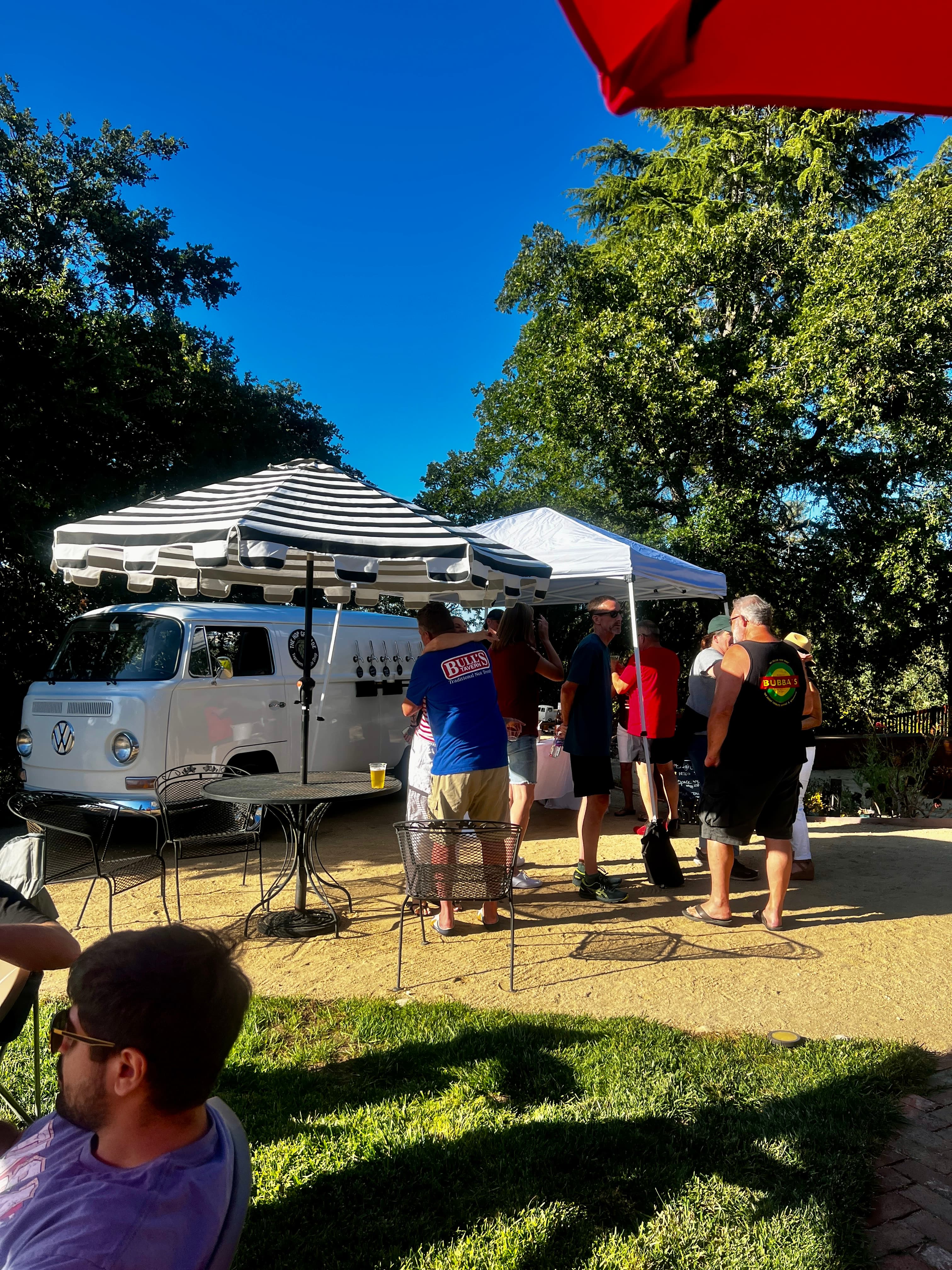 People socialize outdoors near a white vintage VW bus mobile bar and striped umbrella.