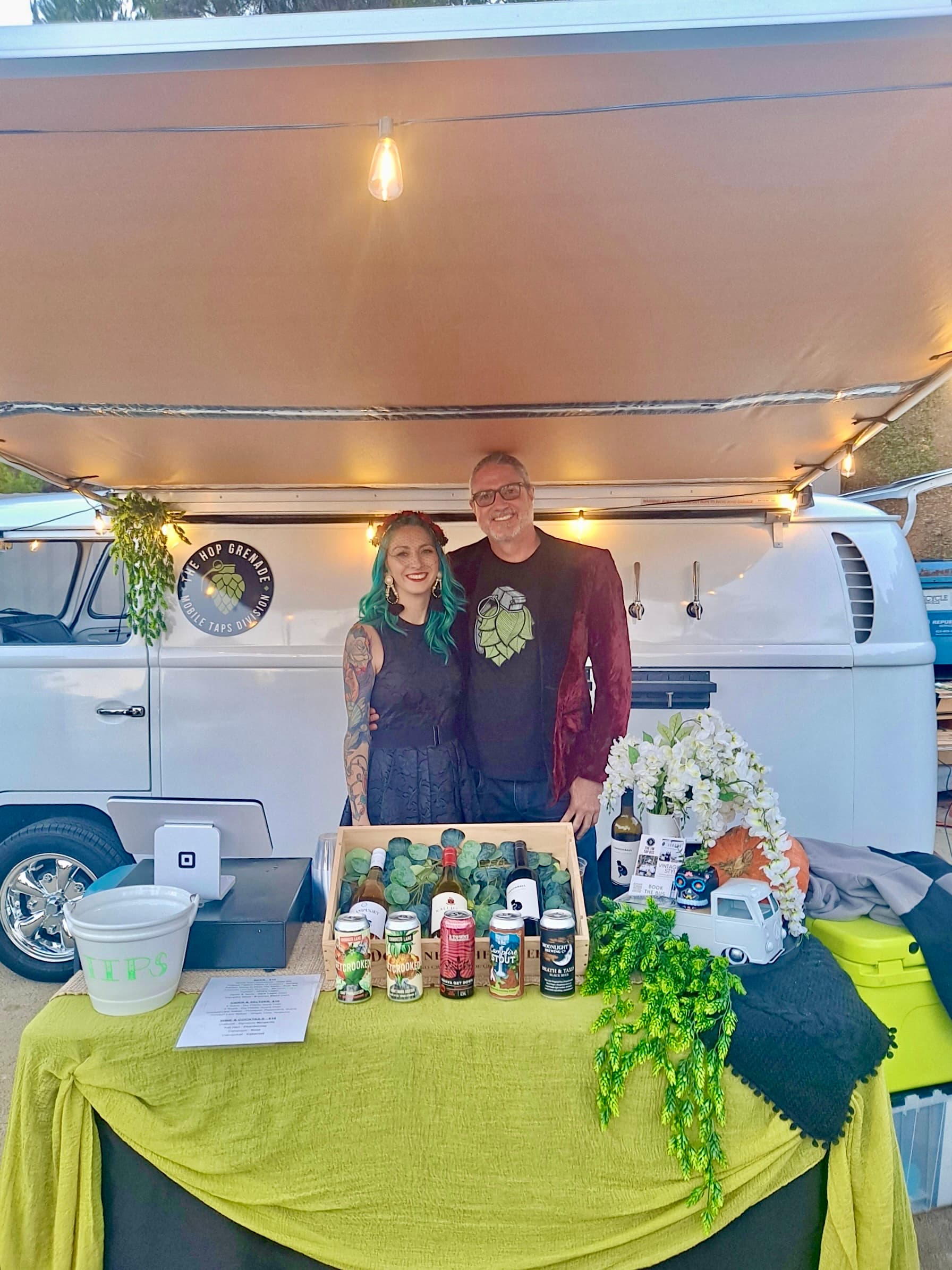 A man and teal-haired woman pose behind a mobile bar and white vintage tap van.