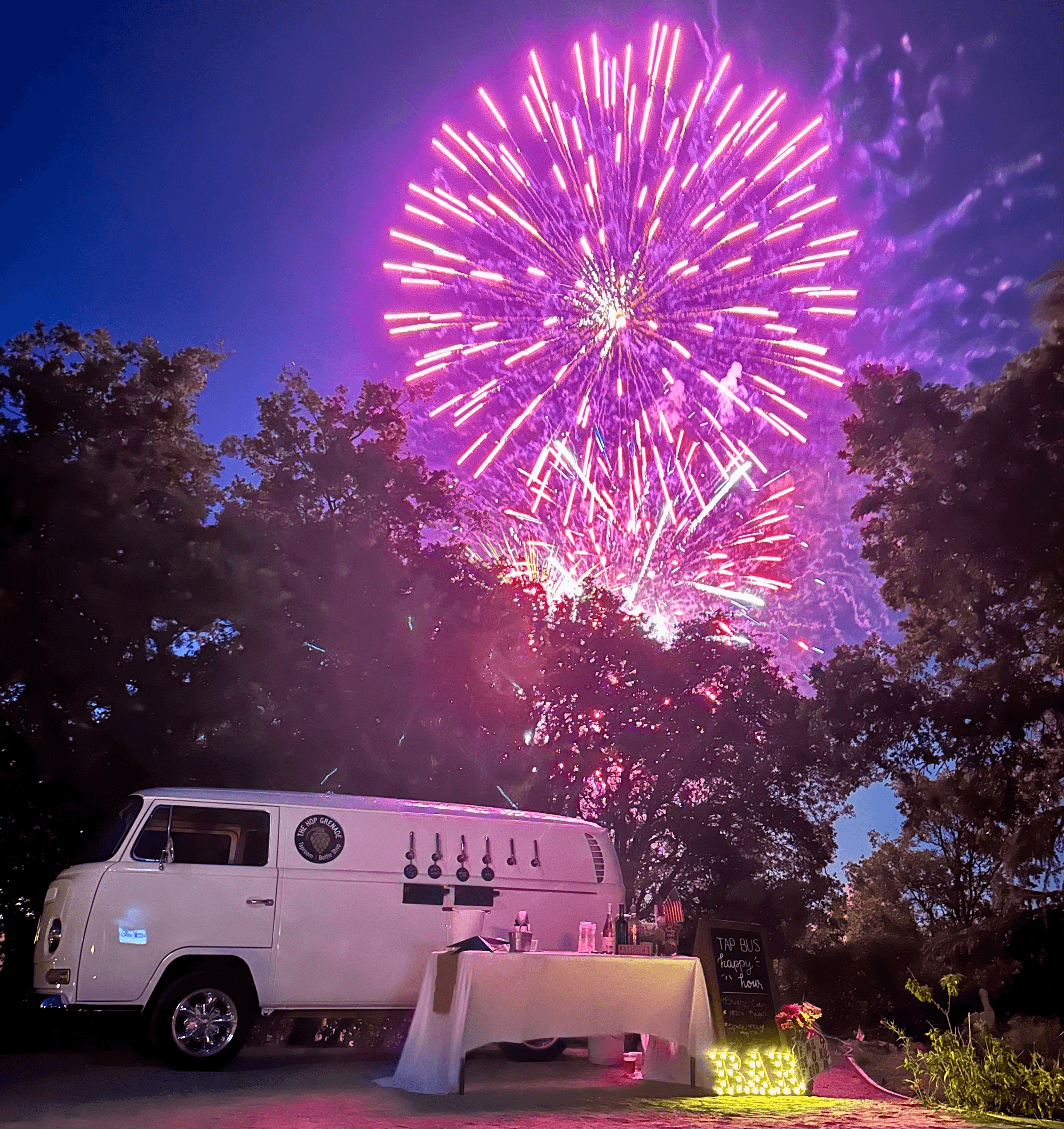 Vibrant purple fireworks explode in the night sky above a white vintage mobile tap bus.