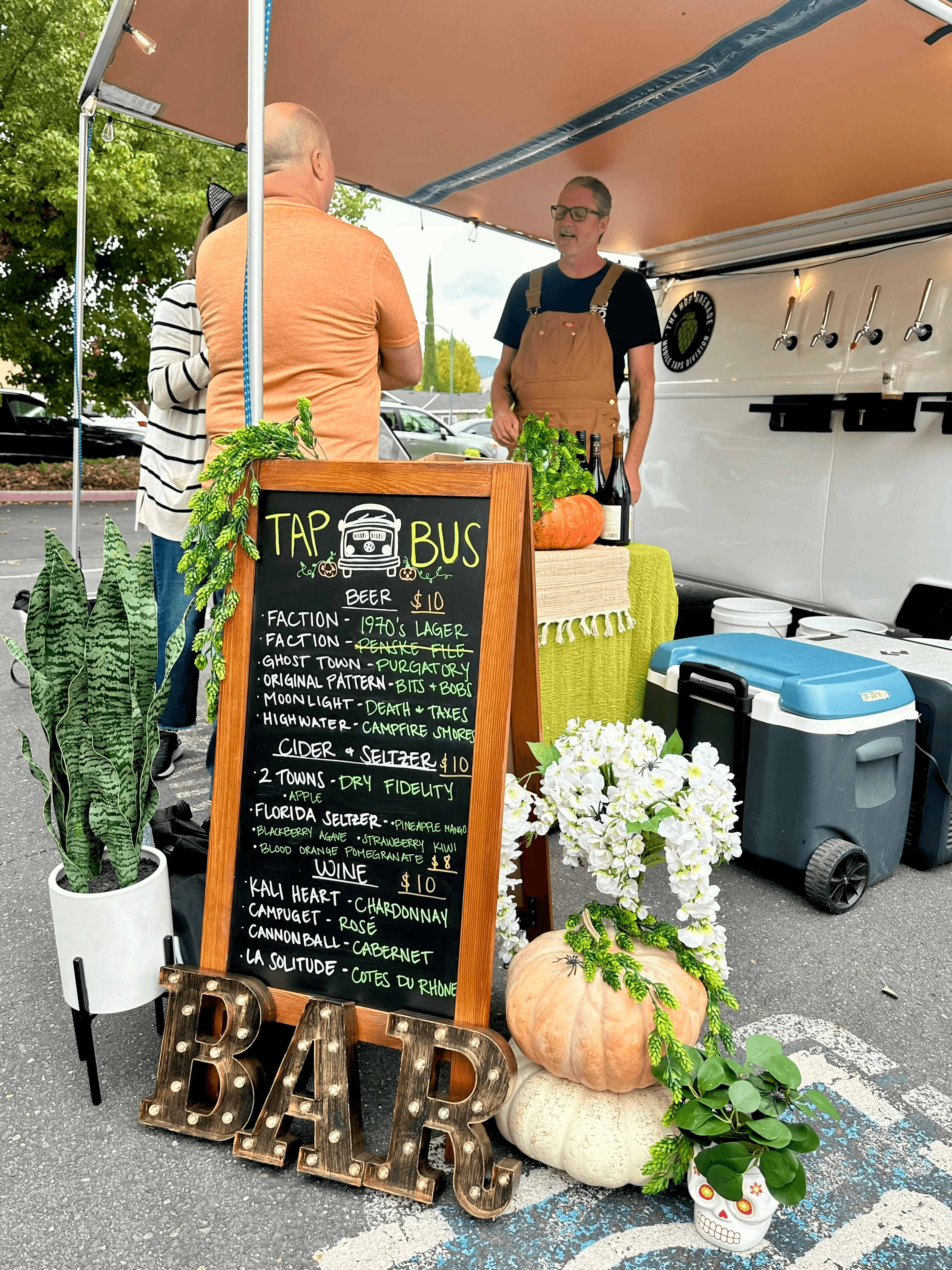 Mobile bar featuring a chalkboard menu, pumpkins, and a bartender serving from a white van.