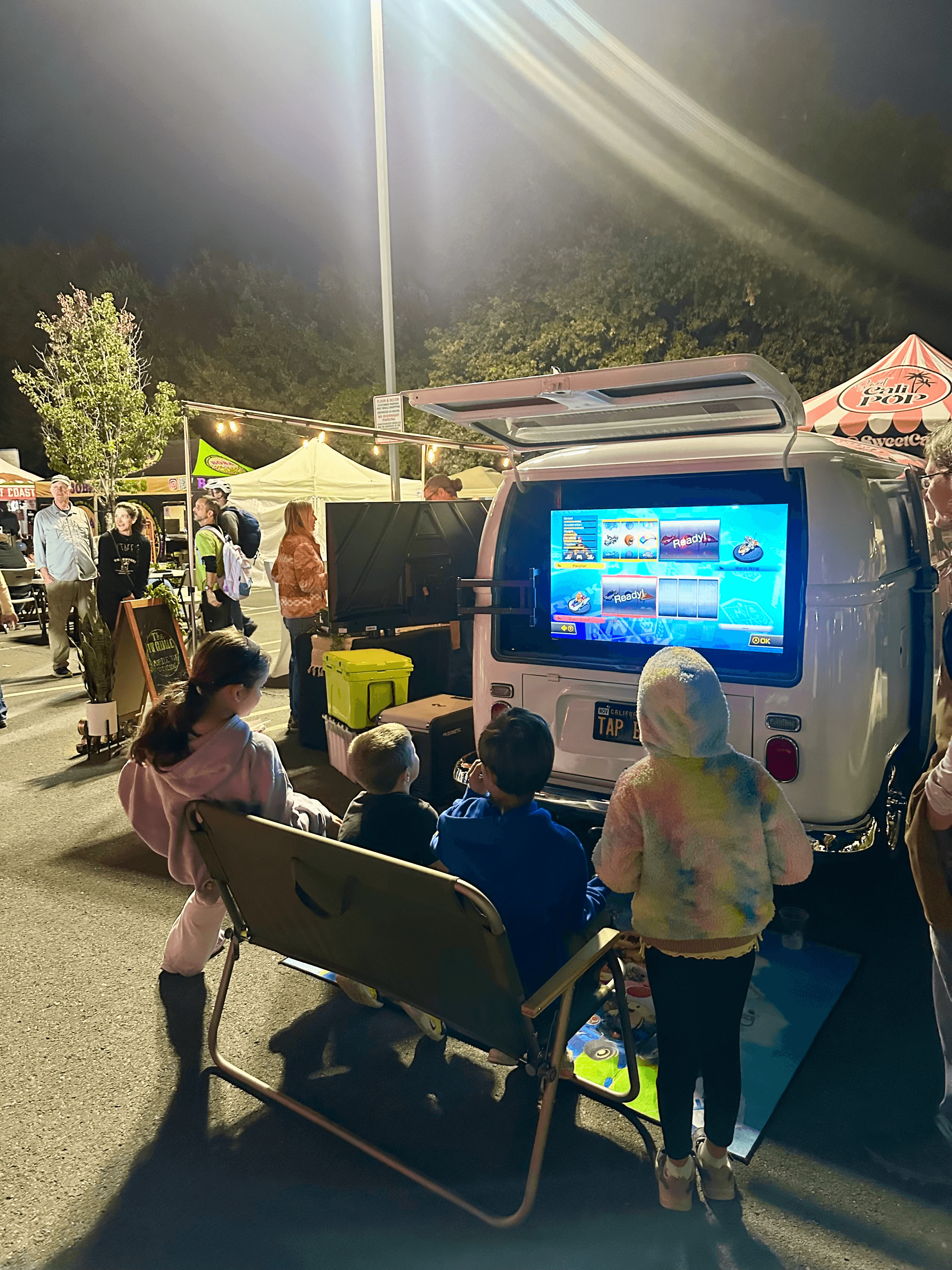 Children play Mario Kart on a screen inside a white van at a night market.