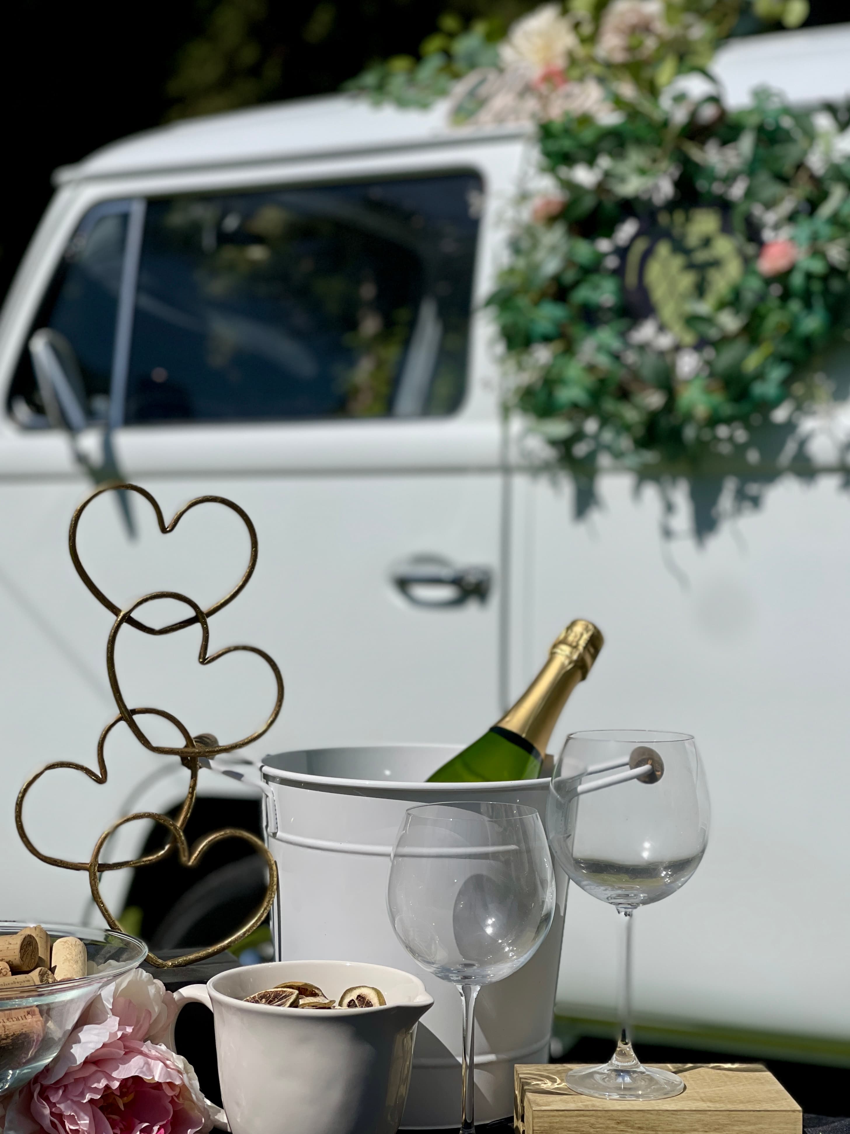 Champagne, wine glasses, and heart sculpture set before a white van with floral wreath.